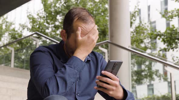 A Caucasian Man Acts Unhappy and Shakes His Head As He Looks at a Smartphone with a Smile