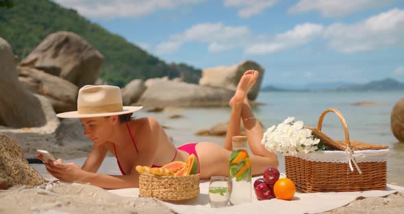 Lovely Woman in Bikini and Hat is Lying with Legs Raised and Texting on a Picnic on Isolated Beach alt