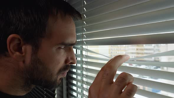A man with a beard approaches the window, pulls back the blinds, and looks out the window through alt