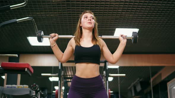 Front View of a Young Woman in Short Top and Leggins in the Gym Doing Squats with a Barbell alt