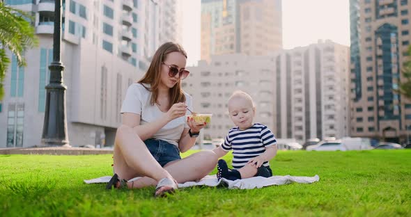 Young Mom with Baby Sitting on the Grass in the Park Eating Lunch alt