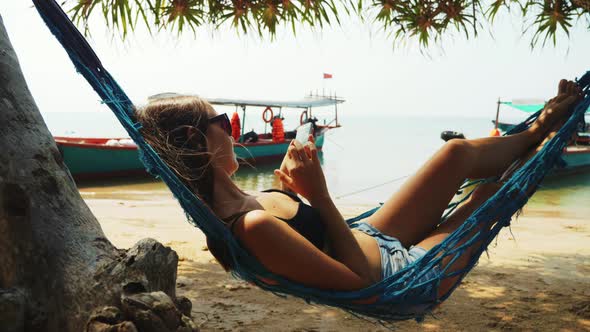 Woman is Resting in Hammock Browsing Pages in Smartphone in Ocean Sand Beach