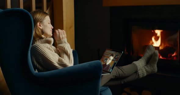 Woman Talks Online with Man By Video on Laptop in Cozy Home with Fireplace