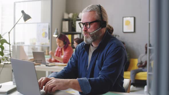 Senior Man in Headset Using Laptop and Talking on Web Call in Call Center alt