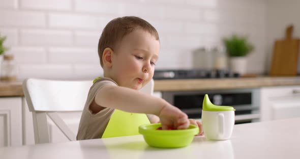 Cute Baby Boy Sits at Table in the Kitchen and Eats Berries with His Hand alt