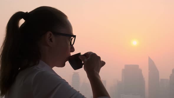 woman drinks tasty coffee while looking out window at evening city at sunset alt