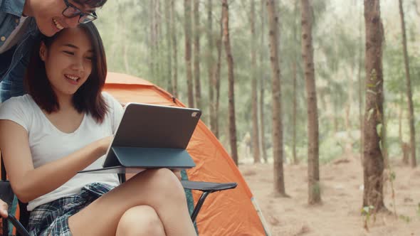 Young asia campers couple using tablet in chairs by tent in forest.