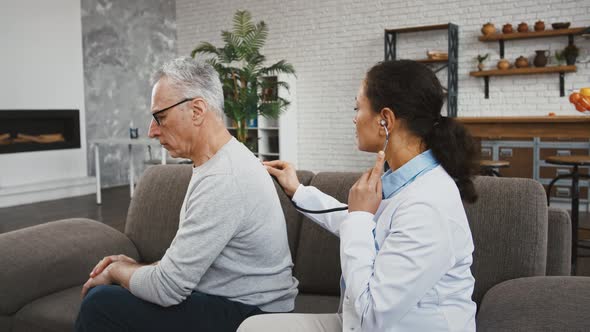 Female Doctor Examining Elderly Male By Phonendoscope Listening to His Lungs Sitting on Couch During alt