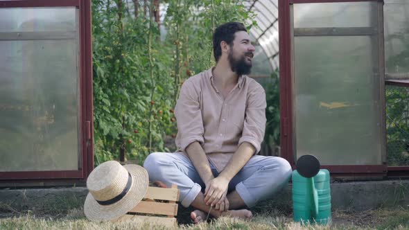Satisfied Happy Smiling Male Farmer Sitting at Greenhouse Entrance Stretching Looking Around alt