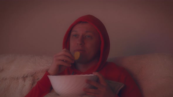 Man in the Hood at Night Watching an Interesting Movie and Eating Chips From a Large Bowl