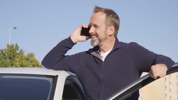 A Middleaged Handsome Caucasian Man Talks on a Smartphone with a Smile As He Stands Next to His Car