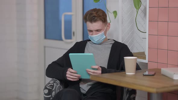 Portrait of Young Positive Man in Covid Face Mask Surfing Social Media on Tablet Smiling alt