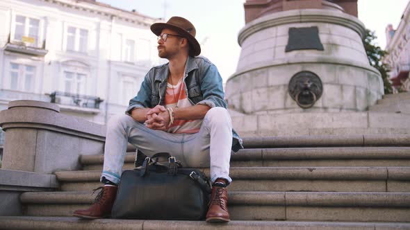 Young Stylish Handsome Redbeard Man with Bag and in Hat Sitting on Stairs in Old City Center alt