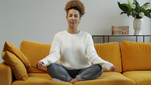 An AfricanAmerican Woman Sitting on a Comfortable Sofa is Doing Yoga in the Lotus Position alt