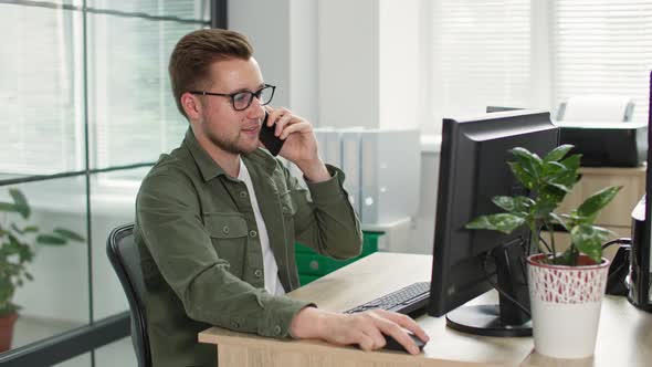 Young Businessman with Glasses Talking on Phone While Working at Computer in Office Background of alt