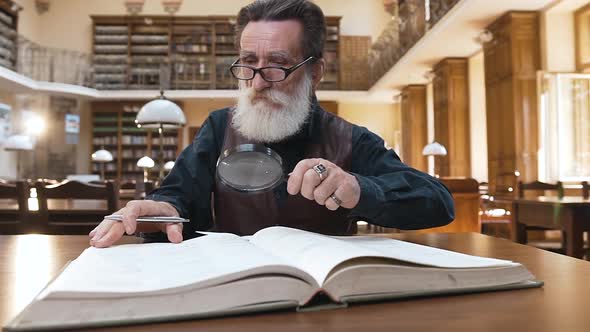Wise Male Pensioner with Beard in Glasses Sitting in the Library and Flipping Through Book Pages alt