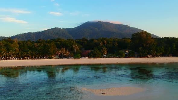Distance and aerial footage of a white sand beach with green and mountain in the background alt