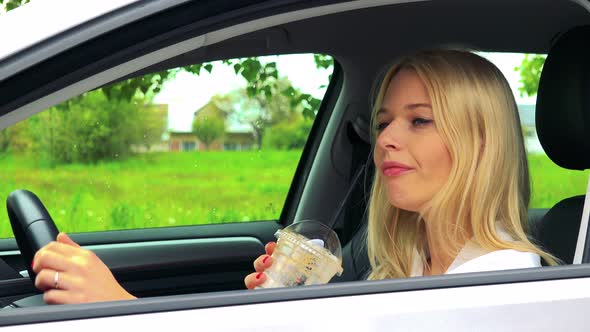Young Attractive Blond Woman Sits in the Car and Drinks Ice Coffee in Cup alt