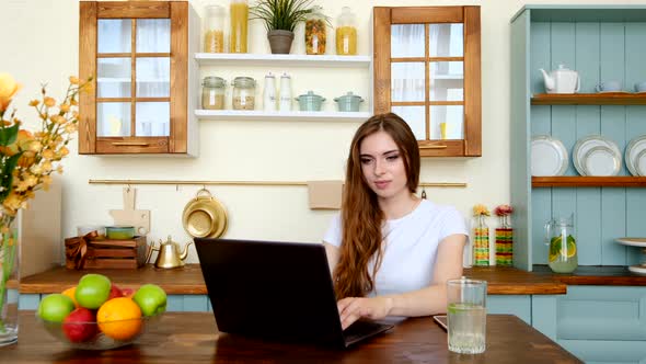 Young Attractive Woman With Dark Hair Sitting At The Table In The Kitchen With Laptop And Working