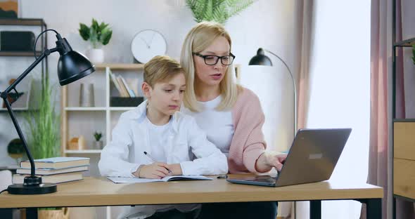 Mother in Glasses which Sitting at Home Desk with Her Handsome Son and Helping Him alt