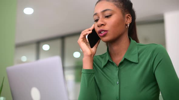 Charming Young AfricanAmerican Woman Using Laptop on the Workplace and Has Phone Conversation
