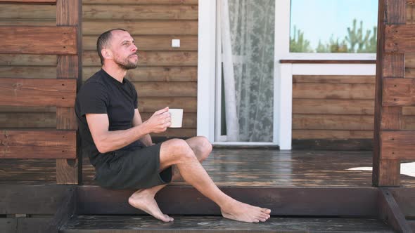 A Man in Shorts Drinks Tea Sitting on the Terrace and Enjoys Nature The Terrace at a Beautiful House