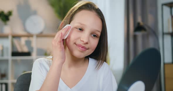 Teen Girl with Long Hair Looking Into Mirror while Uses Facial Cosmetic with Cotton Pad alt