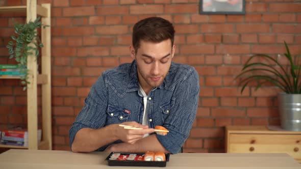 Front View of Happy Young Caucasian Man Sitting in Living Room at Home and Eating Sushi Rolls with alt