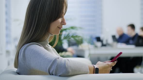 Closeup of a Modern Girl Uses a Mobile Phone on the Couch in the Office Scrolls Types Text Surfs the alt