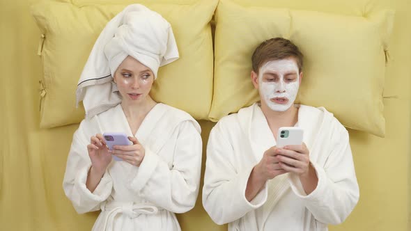 Young Couple in Bathrobes Lying on Bed Using Smartphones Suspecting Each Other
