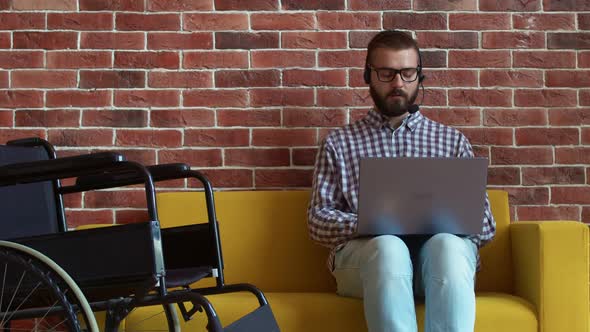 Disabled Person is Sitting on Couch with Laptop in Headset and Talking to Someone Working in Support alt