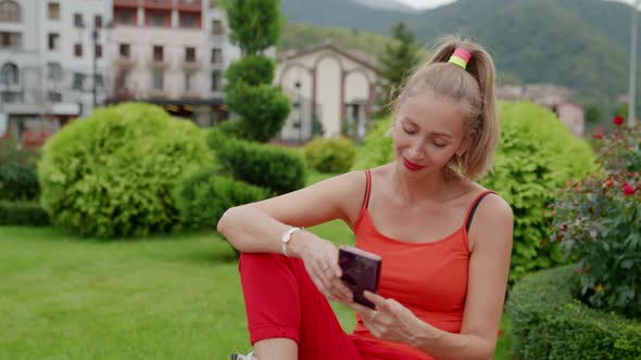 a Blonde in Red Clothes is Sitting with a Phone in Hands Against the Background of Greenery a Hotel alt