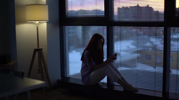 Happy Redhead Young Woman Dials Number and Speaks to Boyfriend Sitting on Windowsill in Evening alt