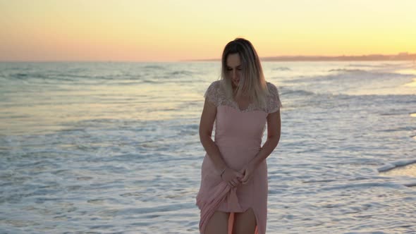 A Young Beautiful Blond Woman in a Pink Dress Walks Along the Beach at Sunset alt