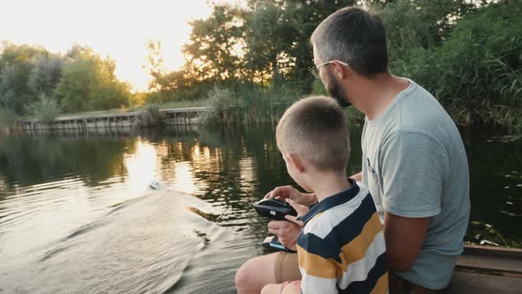 Father and Little Son Play with RC Toy Speedboat on the Lake alt