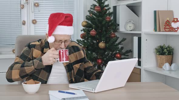 A Middleaged Man in a Santa Claus Hat Drinks a Hot Drink From a Cup and Eats Cookies While Sitting