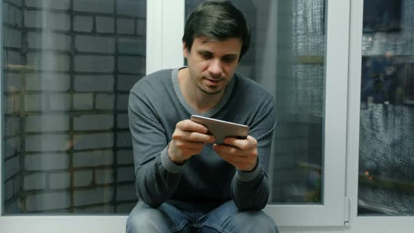 Man Is Playing Games on a Mobile Phone Sitting on Windowsill.