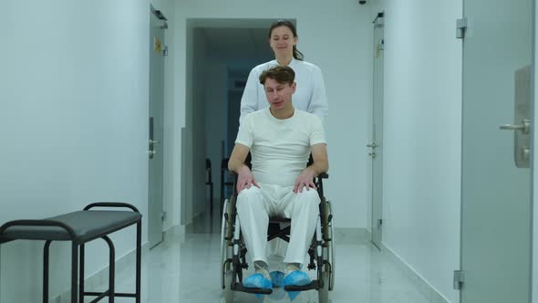Wide Shot Portrait of Positive Doctor Talking to Patient Pushing Wheelchair in Hospital Hallway alt