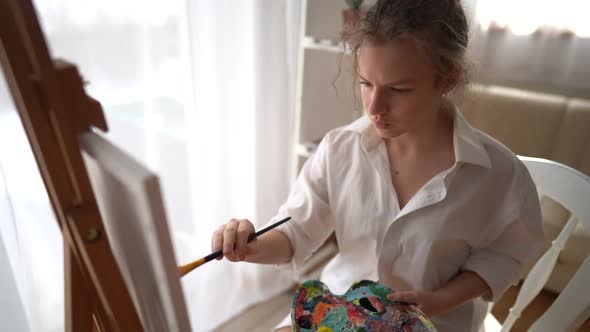 Pretty Girl in a White Shirt with Brush in Hand Sits Near an Easel and Draws on Canvas alt