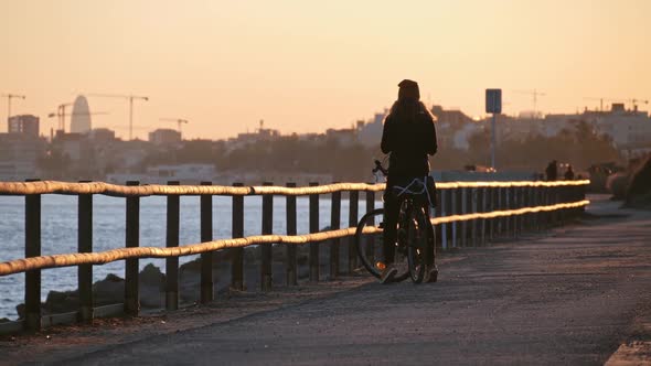 Girl with a Bicycle Standing Close the Sea at the Promenade Road to Barcelona at the Sunset