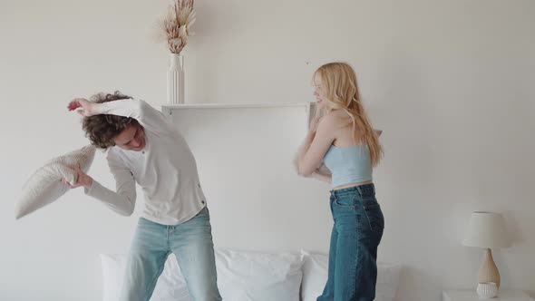Curly Guy and Young Blonde Play Fighting With Pillows Standing On Bed in Modern Interior alt