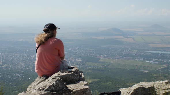 A girl in an orange T-shirt sits on the edge of a cliff, looking out over the city and fields alt