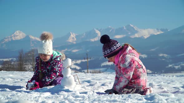 Children in Outwear Making Small Snowman While Playing on Snowy Field in Sunlight with Mountains on