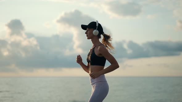 A Young Woman Runner is Listening to Music in Earphones and Training By a Sea alt