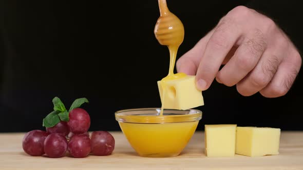 Man tasting cheese with honey and grapes. Dips wooden stick to glass bowl with liquid honey alt