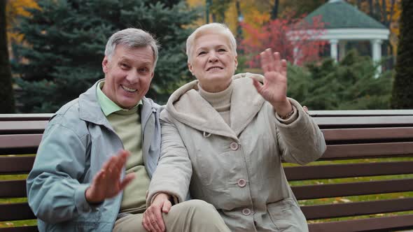 Caucasian Family Couple Lovely Elderly Married Grandparents Sit on Bench in Park Wave Hands Welcome alt