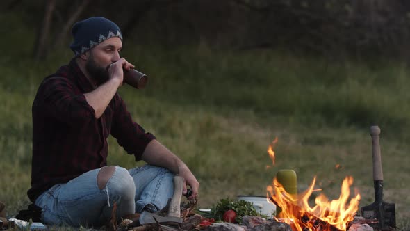 Thoughtful Man Tourist Sits By Campfire and Looks Into the Blazing Fire Drinks From Bottle