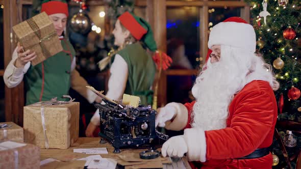 Santa and Elves with presents on Christmas decor indoor. Santa Claus is typing letter alt