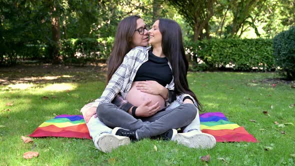 Portrait of Affectionate Pregnant Lesbian Couple with Rainbow Flag Relaxed at the Park alt