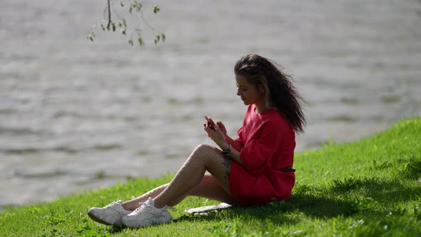 Young Lady with Smartphone on Shore of River in City at Summer Day Internet Addiction alt